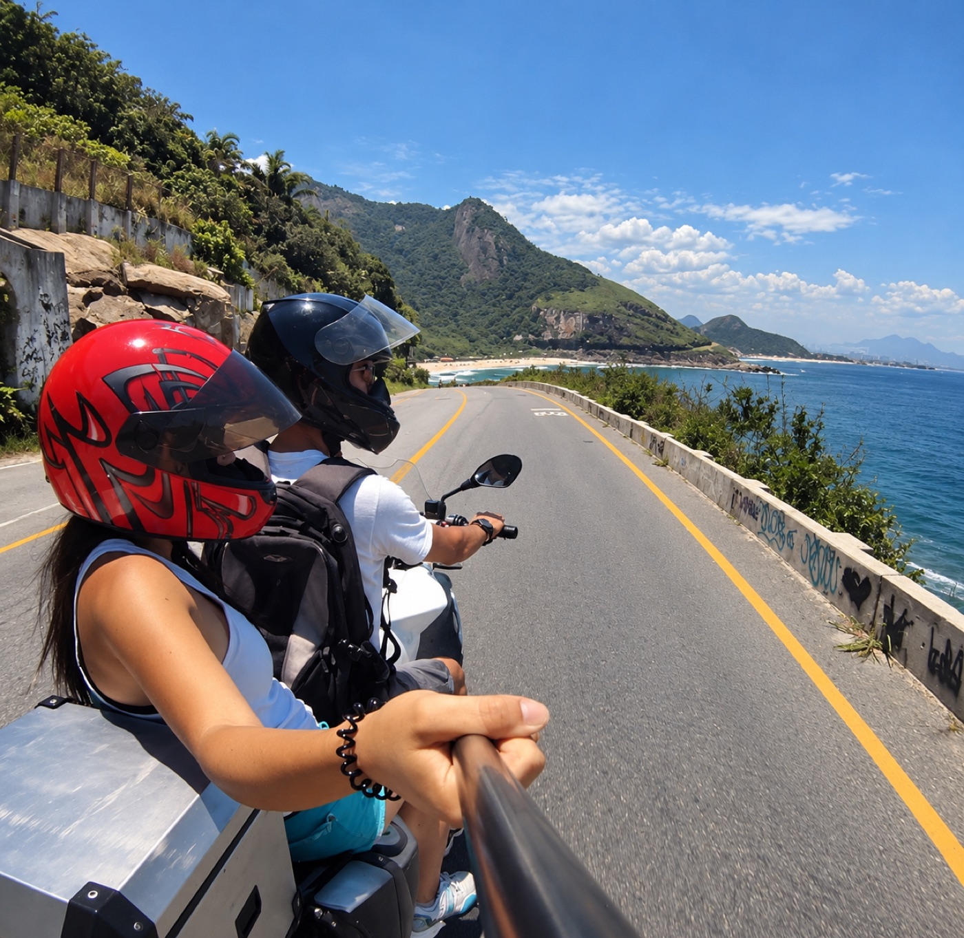 Beifahrerin mit rotem Helm auf der Ozeanstraße in Rio, vor ihr Strand und Berge